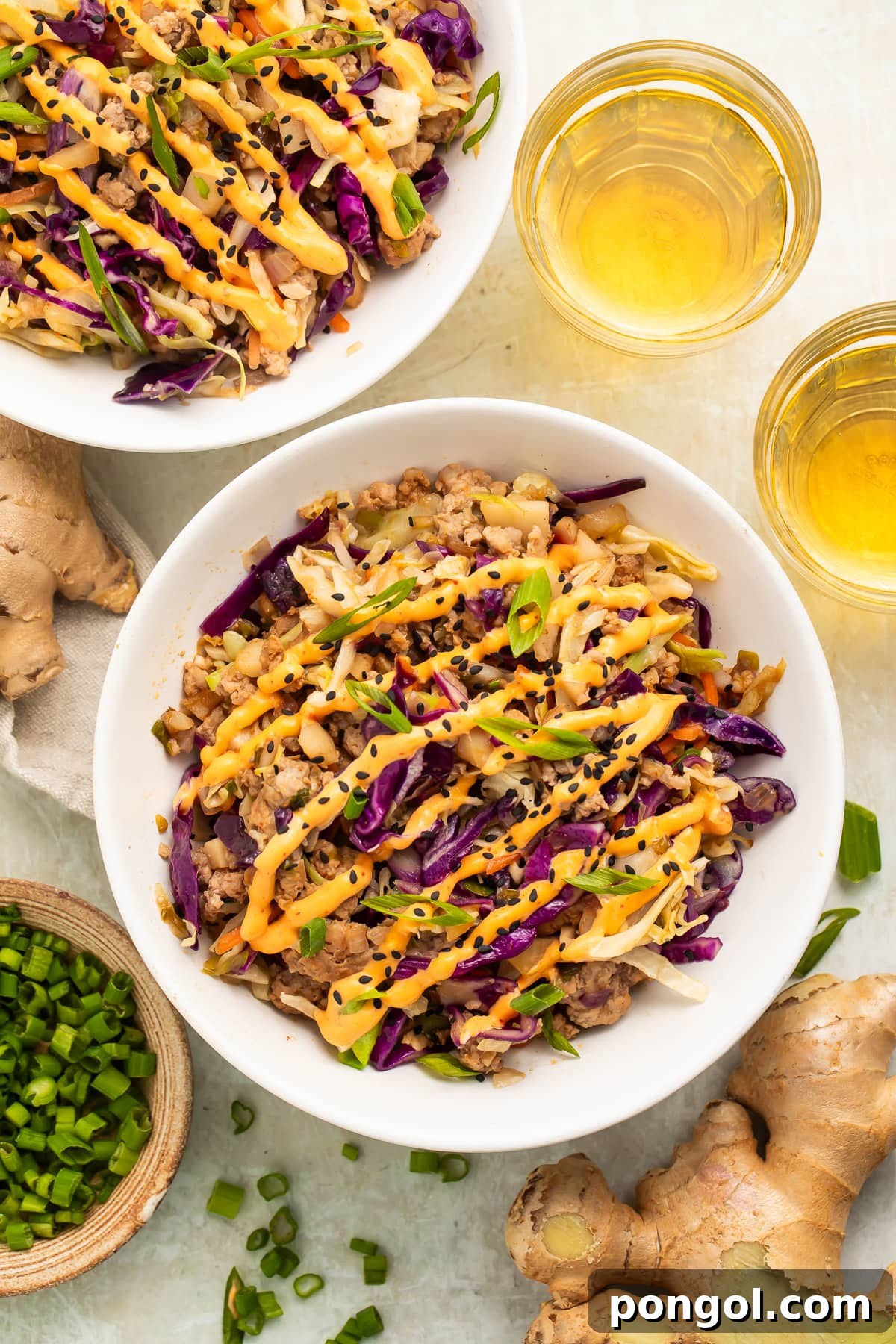 View of egg roll bowl with ground pork and vegetables topped with orange sauce, beverages visible in background