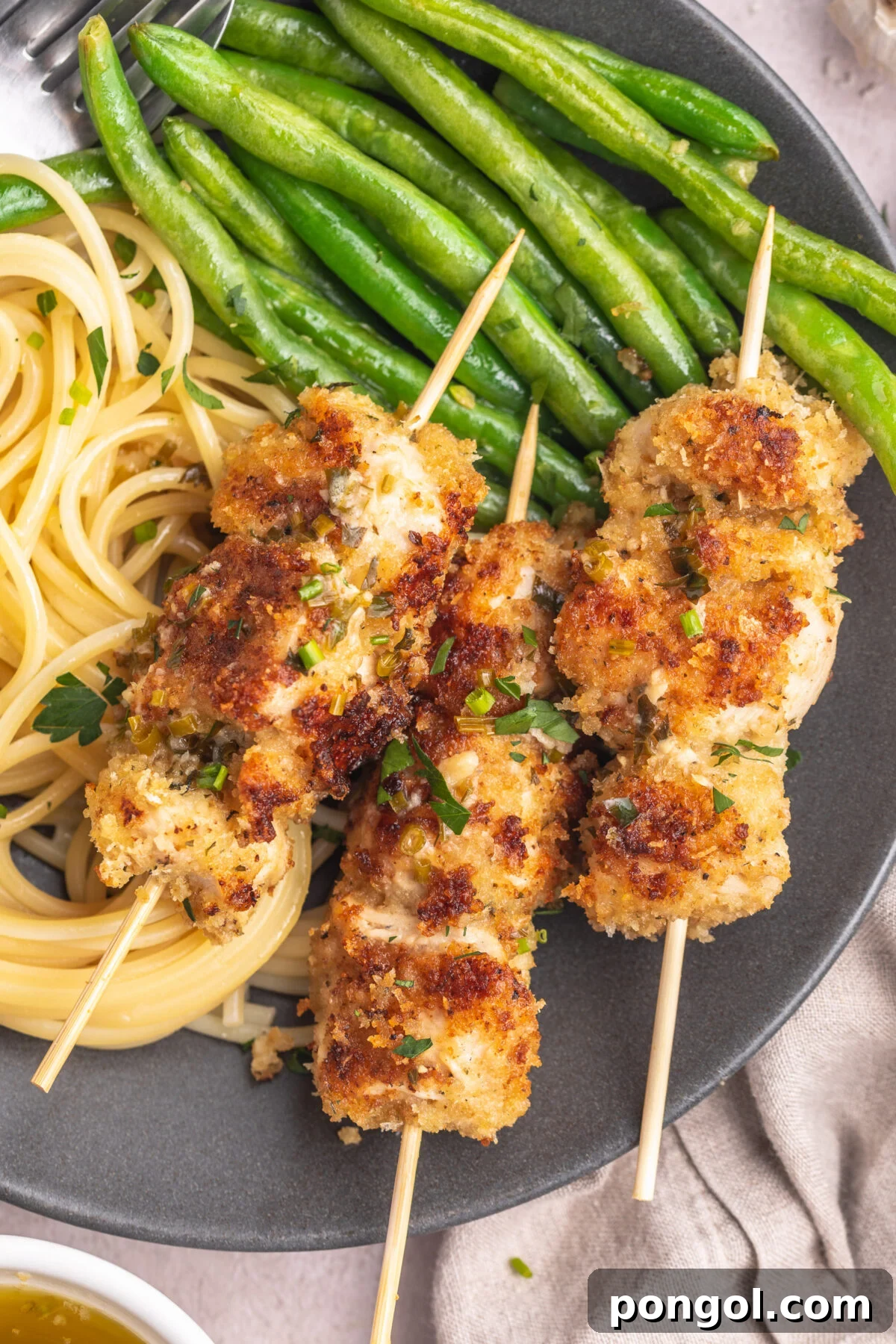 Three chicken spiedini skewers on a dark grey plate with angel hair pasta and bright green beans topped with an herb butter sauce.