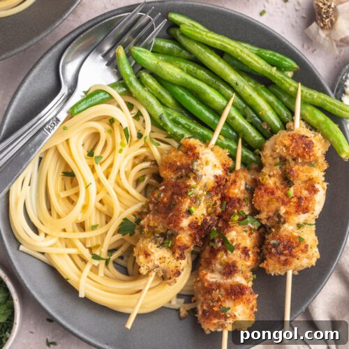 Top down view of three chicken spiedini skewers plated on a dark grey plate next to a small portion of angel hair pasta and a pile of bright green beans.