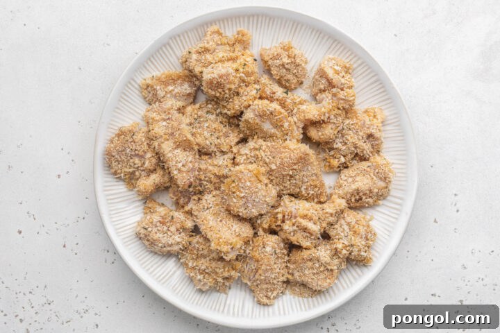 Breaded boneless, skinless chicken breast chunks on a large white plate ahead of being skewered.