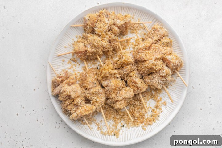 Breaded boneless, skinless chicken breast pieces threaded onto wooden skewers and stacked on a white plate.