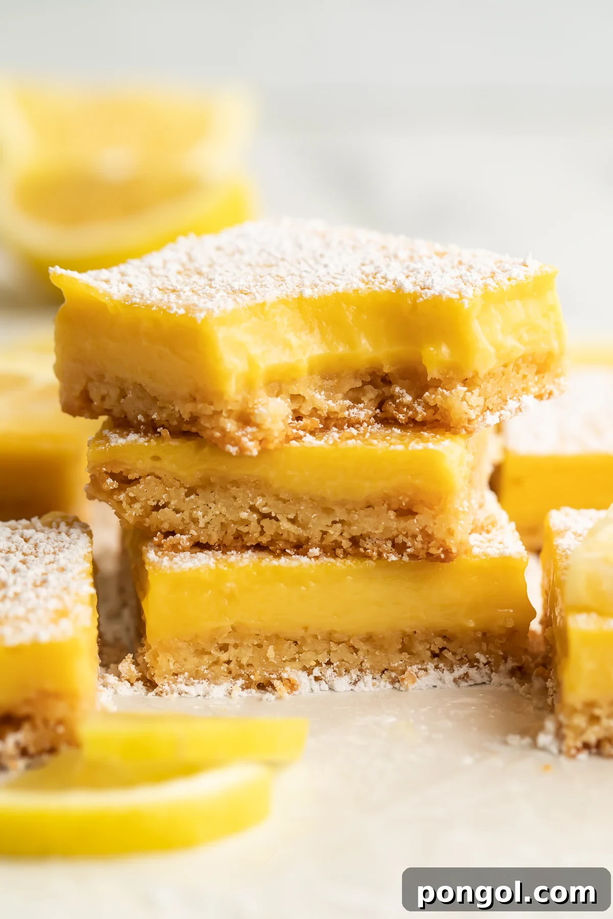 Three paleo lemon bars stacked on top of each other. A bite is missing from the corner of the lemon bar on top. Other lemon bars surround the stack, just out of focus.