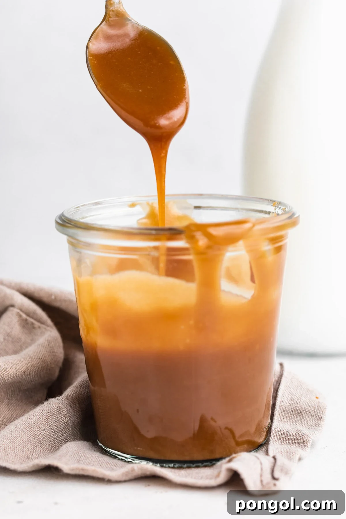 Decadent 3-Ingredient Keto Caramel 2 Side view of a jar of keto caramel sauce. A silver spoon hovers above the glass jar, drizzling caramel back in to the jar.