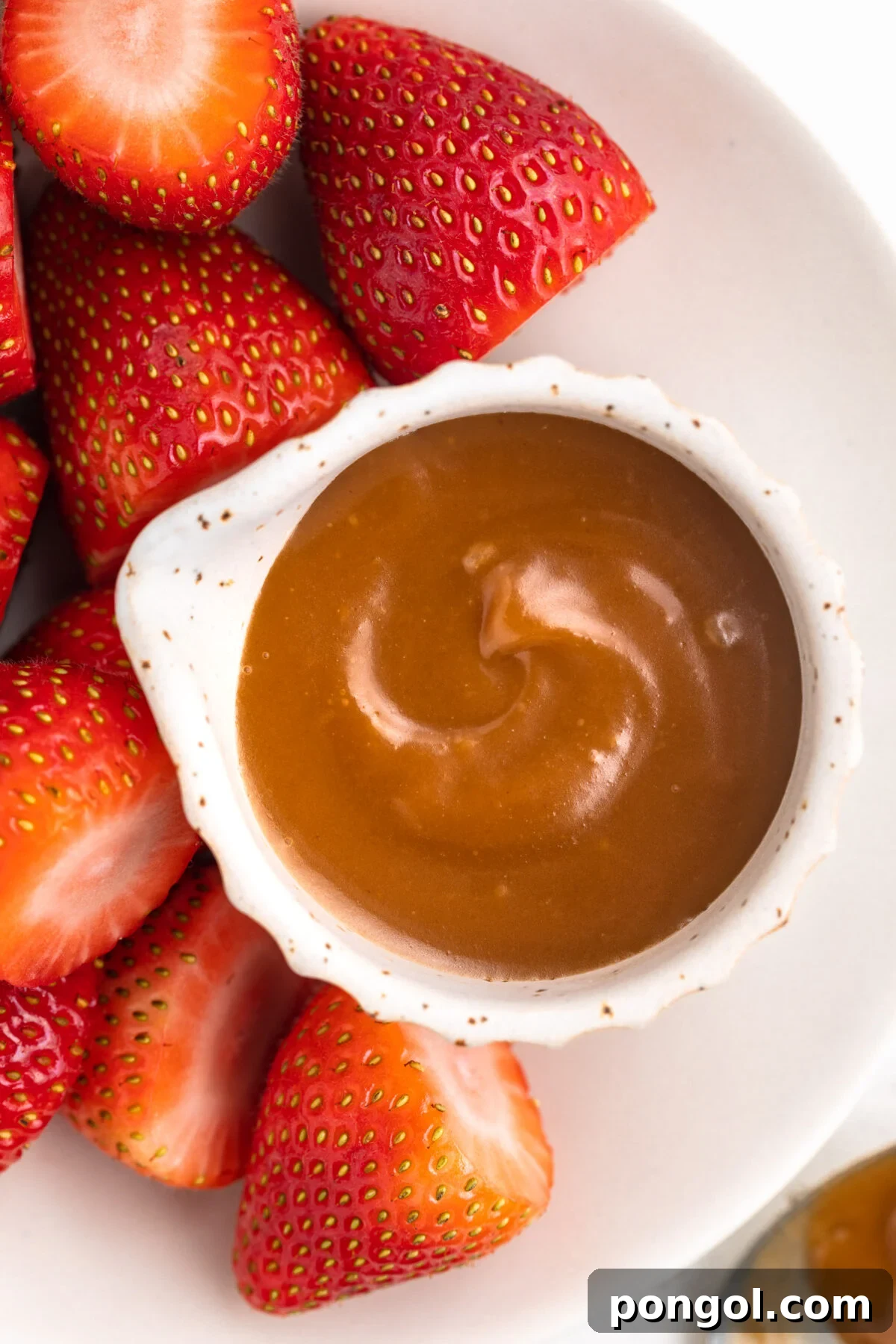 Decadent 3-Ingredient Keto Caramel 3 Overhead look at a small ramekin of keto caramel sauce on a plate next to sliced bright red strawberries.