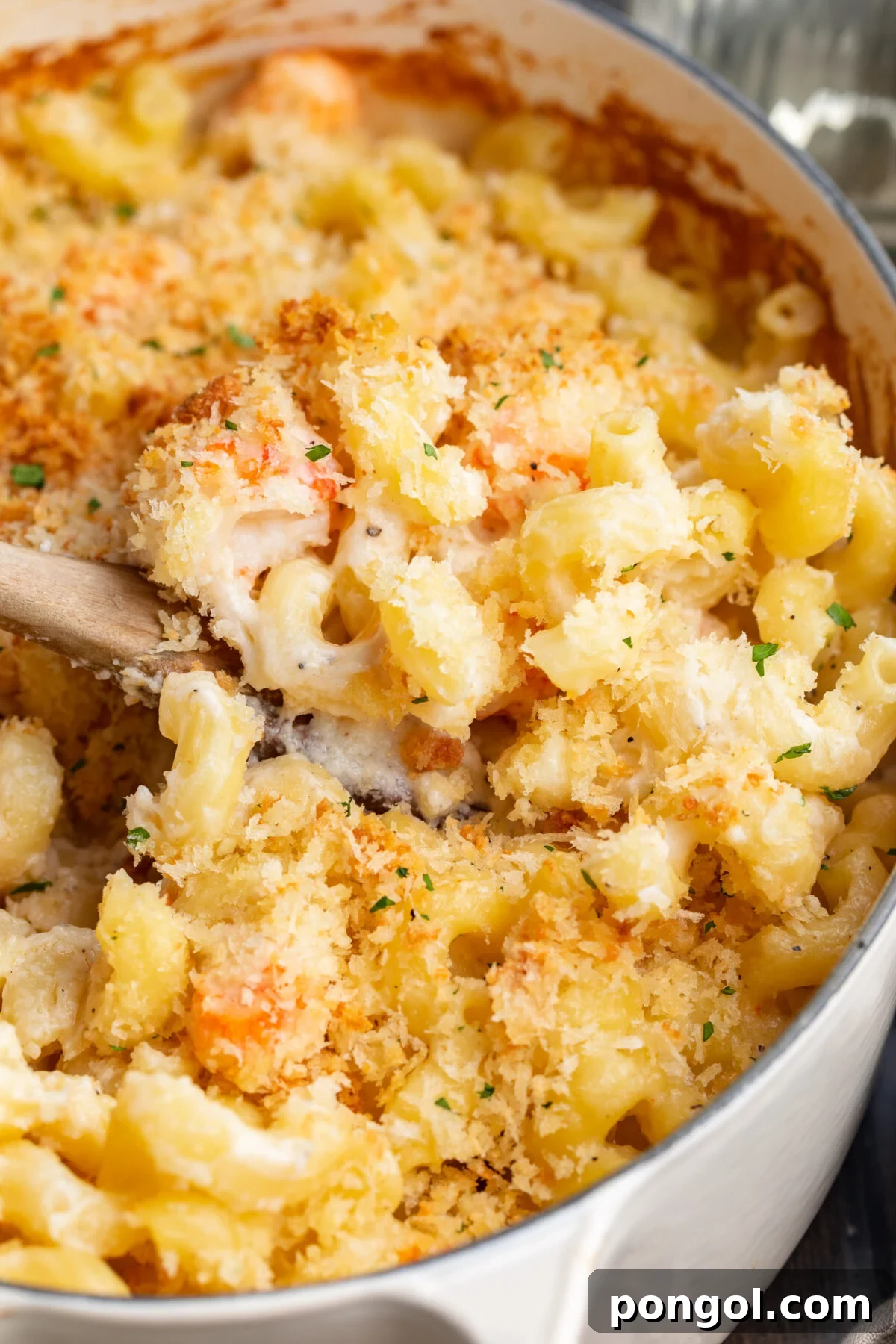 Decadent Lobster Mac 3 Close-up view of a scoop of lobster mac and cheese resting on a wooden spoon being lifted out of a casserole dish.