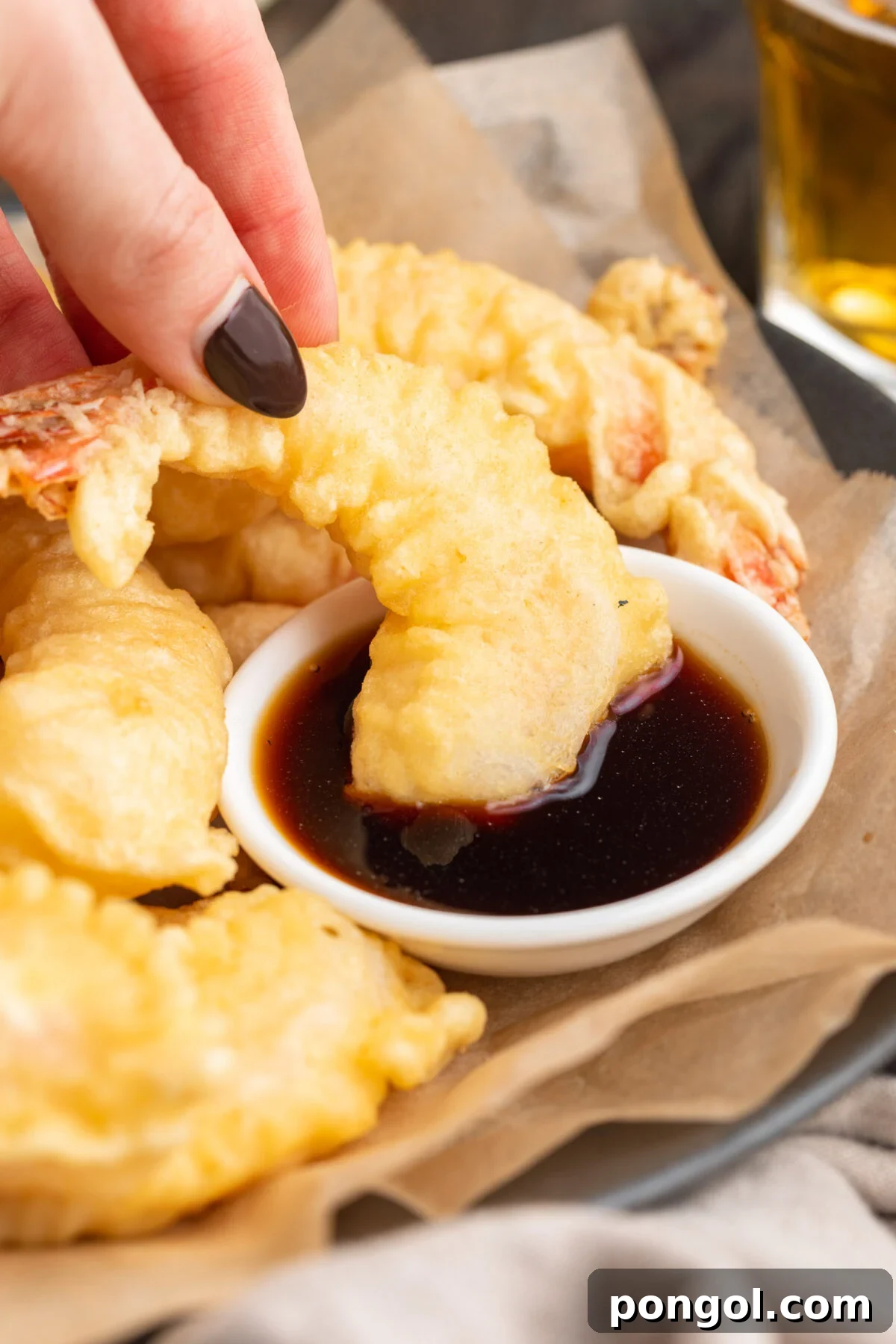 Golden Crispy Shrimp Tempura 3 A close-up view of a hand dipping a perfectly golden-brown and crispy shrimp tempura into a small ceramic bowl filled with a dark, savory soy-based dipping sauce.