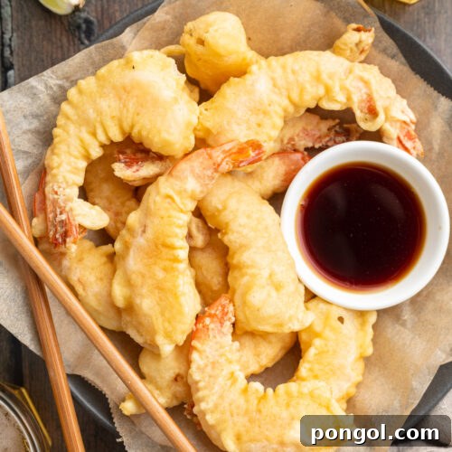 Zoomed-out, overhead photo of perfectly fried shrimp tempura arranged neatly on parchment paper next to a small bowl of savory tempura dipping sauce, with chopsticks in anticipation of serving.