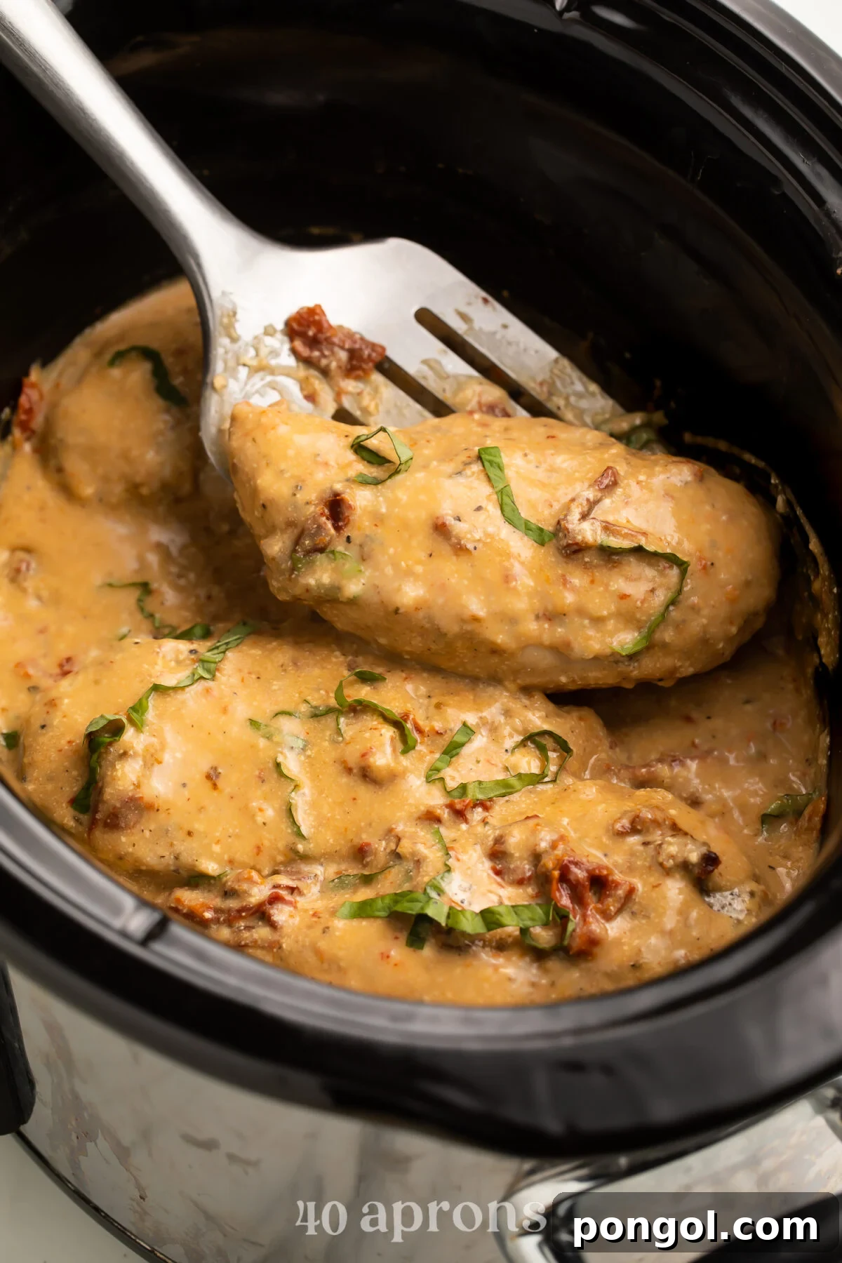 A Marry Me Chicken breast being lifted out of a Crockpot of sauce with a silver spatula.