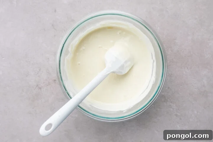 Melted white chocolate and coconut oil in a large glass mixing bowl with a white silicone spatula.