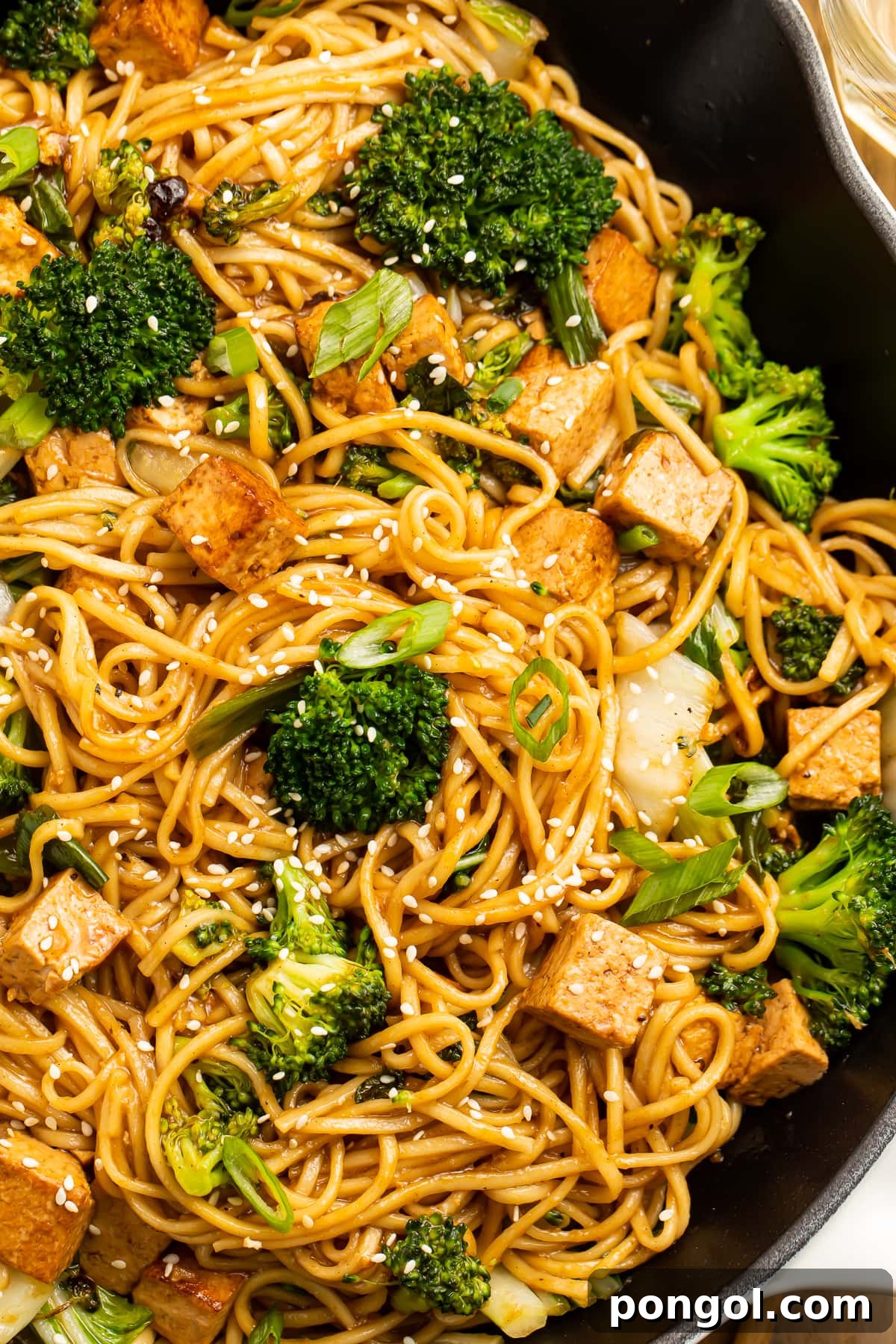 Close-up view of stir fry noodles with tofu and vibrant vegetables in a cast iron skillet, garnished with sesame seeds and green onions.