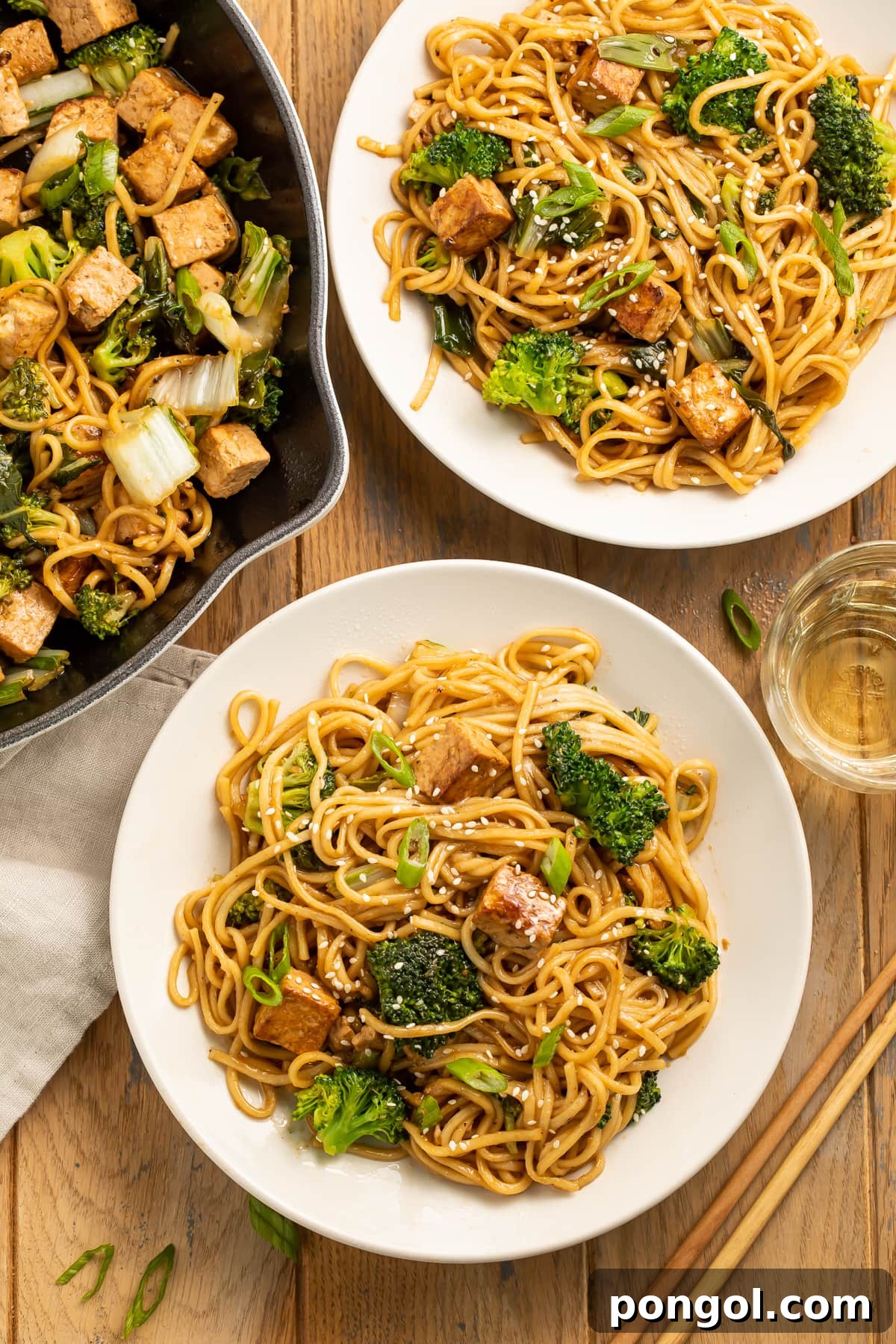 Overhead view of two large white plates piled with stir fry noodles and tofu next to a skillet of noodles, beautifully plated and ready to serve.